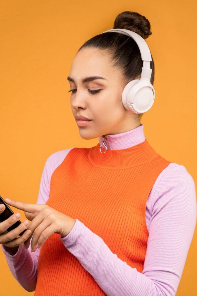 Woman with white headphones using smartphone against an orange background.